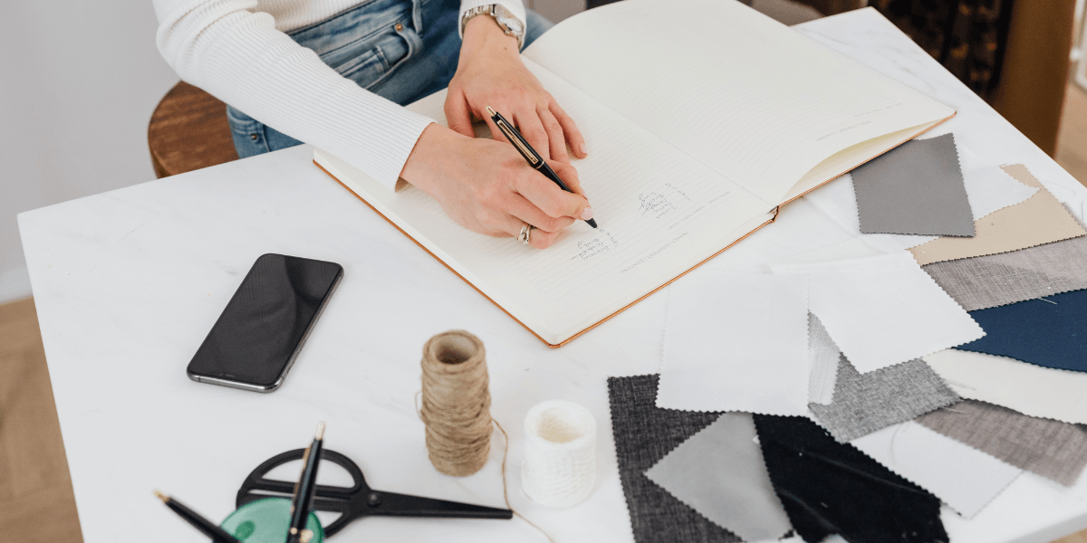 Designer writing in a notebook with fabric swatches and tools spread across a white desk.